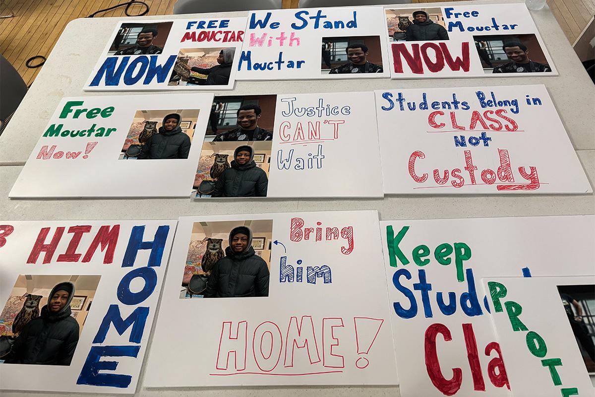 A table full of signs created by Brooklyn Frontiers High School staff: "Free Mouctar Now," "We Stand with Mouctar," "Justice Can't Wait," "Students Belong in CLASS Not in Custody," "Bring Him Home!"
