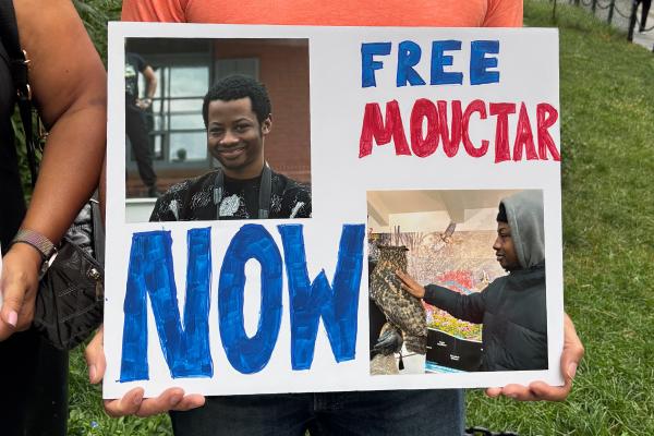 Close up of a hand made sign "FREE MOUCTAR NOW" with photos of a smiling young African man.