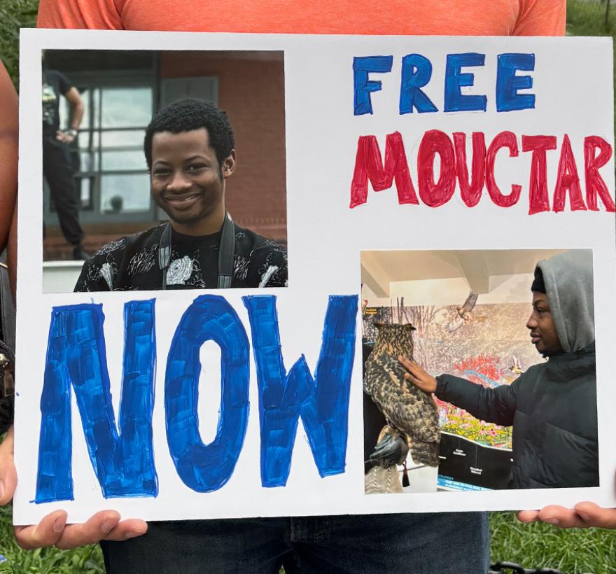 Close up of a hand made sign "FREE MOUCTAR NOW" with photos of a smiling young African man.