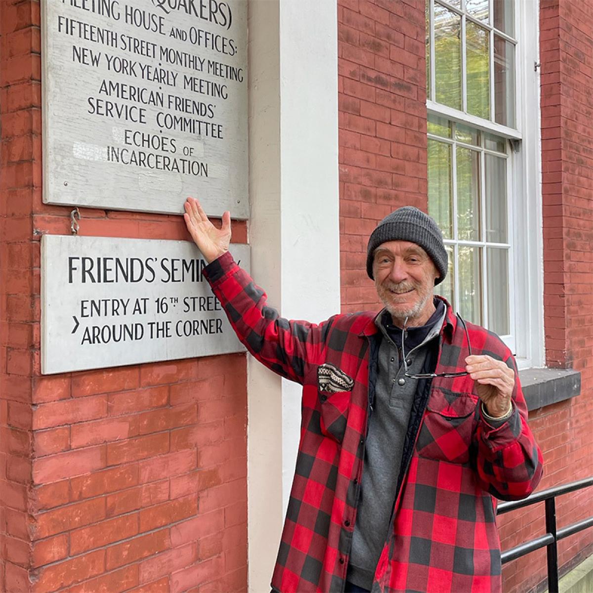 An older white man (John Maynard) wearing a red flannel shirt is smiling and pointing to a sign on the front of the 15 Rutherford Place building that he has just touched up with paint. From November 2023.